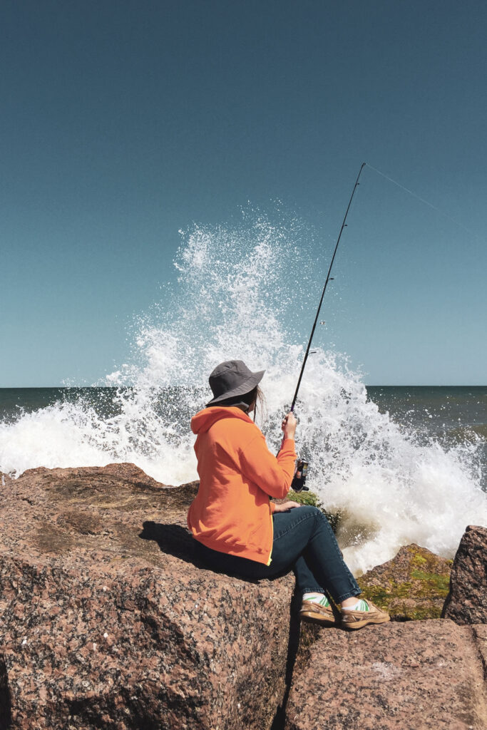 Alyx fishing in a bright orange jacket with a wave hitting the rocks